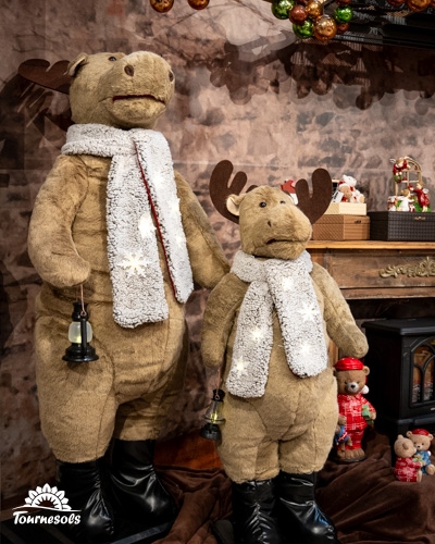 Deux rennes en peluche portant des écharpes blanches, entourés de décorations de Noël.